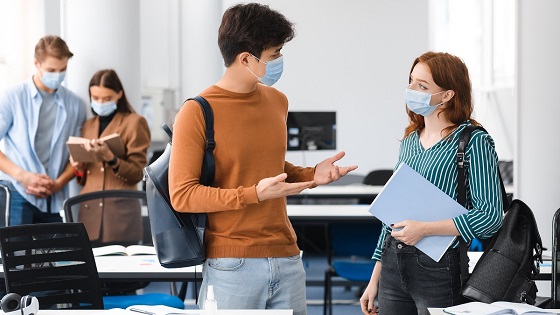 Two university students standing in classroom with face masks on, discussing